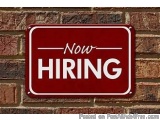 The image is a rectangular red sign with white text that reads "Now Hiring". The sign is mounted on a brick wall. The text is...