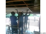 The image shows three men working on a wooden structure in a garage or workshop. They are standing on ladders and are holding...