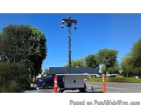 The image shows a tall pole with multiple lights attached to it. The pole is located in a parking lot with trees and bushes i...