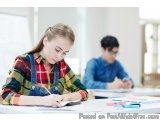 The image shows a young woman sitting at a desk in a classroom. She is wearing a plaid shirt and denim overalls and is holdin...