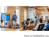 The image shows a man standing in front of a group of people in a conference room. He is holding a microphone and appears to...