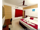 The image shows a hotel room with a large double bed in the center. The bed is covered with white linens and red pillows, and...