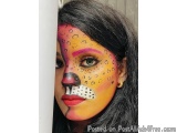 The image is a close-up portrait of a woman's face. She has long dark hair and is wearing a colorful face paint design. The d...