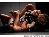 The image shows two men engaged in a mixed martial arts (MMA) fight. The man on the left is wearing a white tank top with the...