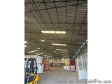 The image shows the interior of a large warehouse with a high ceiling and metal beams. The warehouse appears to be empty, wit...