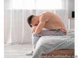 The image shows a young man sitting on a bed with his head in his hands. He is wearing a peach-colored t-shirt and grey strip...