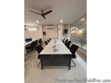 The image shows a modern conference room with a long white table in the center. The table is surrounded by black chairs with...