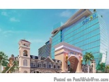 The image is a digital rendering of a modern hotel and casino complex. The complex is located in a tropical location with pal...