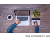 The image shows a person's hands typing on a laptop keyboard on a wooden desk. The laptop is open and the screen is turned on...