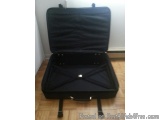 The image shows a black suitcase with its lid open on a wooden floor. The suitcase appears to be made of a hard material and...