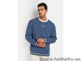 The image shows a young man standing against a plain white background. He is wearing a blue sweatshirt with a white collar an...