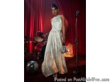 The image shows a young woman standing in front of a red curtain with a drum set in the background. She is wearing a long, fl...
