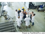 The image shows a group of people in a factory or workshop. They are all wearing white lab coats and hard hats, and are gathe...