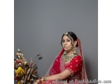 The image is a portrait of a young Indian woman wearing a traditional red and gold lehenga. She is sitting on a chair with he...