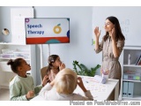 The image shows a classroom with a teacher standing in front of a whiteboard and a group of children sitting around a table....