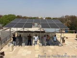 The image shows a group of men standing on a rooftop with a large solar panel installation. The installation is made up of mu...