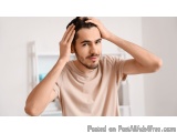The image shows a young man standing in a room with a white wall and a window in the background. He is wearing a light pink t...