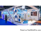 The image shows a trade show booth with a blue and white color scheme. The booth has a modern design with a curved roof and a...