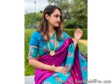 The image is a portrait of a young woman wearing a traditional Indian saree. She is sitting on a stone bench in a garden with...