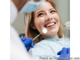 The image shows a young woman sitting in a dental chair with her mouth open, smiling widely. She is wearing a blue dental uni...