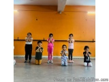 The image shows a group of seven children in a dance studio. They are standing in a line with their hands covering their mout...