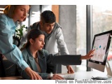 The image shows three people in an office setting, two women and a man, working together on a computer monitor. The woman on...