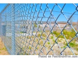 The image shows a chain-link fence made of metal wire mesh. The fence is located in an open area with a clear blue sky in the...