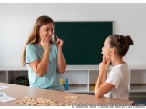 The image shows a young girl and a young woman sitting at a desk in a classroom. The girl is wearing a light blue t-shirt and...