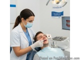 The image shows a young man sitting in a dental chair with his mouth open, while a dentist, wearing a white coat and a face m...