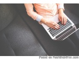 The image shows a person sitting on a gray couch with a laptop on their lap. The person is wearing a pink and white checkered...