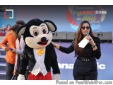 The image shows a woman standing next to a mascot of Mickey Mouse. The mascot is wearing a black suit with a white shirt and...