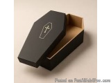 The image shows a black coffin with a gold cross on the lid. The coffin appears to be made of cardboard and has a rectangular...