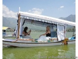 The image shows a traditional boat on a lake with mountains in the background. The boat is white with a blue canopy and has a...