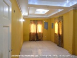 The image shows an empty room with yellow walls and a white ceiling. The room has a tiled floor and a window with sheer curta...