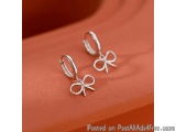 The image shows a pair of silver hoop earrings with a small bow charm hanging from them. The earrings are resting on an orang...