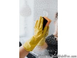 The image shows a person's hand wearing a yellow rubber glove and holding a sponge. The person is cleaning a window with the...