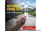The image shows a hand holding a remote control in front of a black metal gate. The gate is located in a residential area wit...