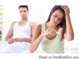 The image shows a young woman in a green tank top sitting on a bed with a man in the background. The woman is holding a pregn...