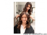 The image shows a young woman sitting in a salon chair with her hair styled by another woman standing behind her. The woman s...