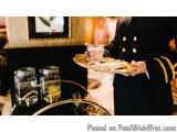 The image shows a person in a black suit with gold buttons and a gold trim, holding a tray with a glass of water and a plate...