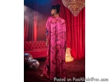 The image shows a woman standing in a dimly lit room with a red curtain in the background. She is wearing a long, flowing pin...