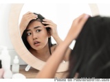 The image shows a young woman looking at her reflection in a round mirror. She is holding her head with both hands and appear...