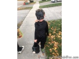 The image shows a young boy dressed up as Batman, standing on a sidewalk with his hands on his chin. He is wearing a black Ba...