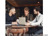 The image shows three men sitting at a wooden table in a cafe or restaurant. They are engaged in a conversation and appear to...