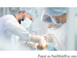 The image shows three surgeons performing an operation on a patient in an operating room. The surgeon on the left is wearing...