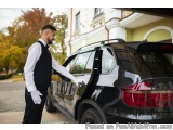 The image shows a man in a black suit and white gloves standing next to a black SUV. He is holding the door open and appears...