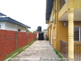 The image shows a narrow street with a yellow building on the right side. The building has a balcony with a railing and a win...
