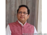 The image is a portrait of a middle-aged man. He is wearing a pink Nehru jacket and a white shirt. He has a pair of glasses o...