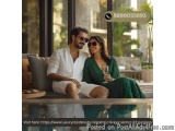 The image shows a young couple sitting on a couch in a living room. The man is wearing a white kurta and shorts, while the wo...