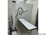 The image shows a medical examination table in a room with a white wall and a door on the right side. The table is made of me...